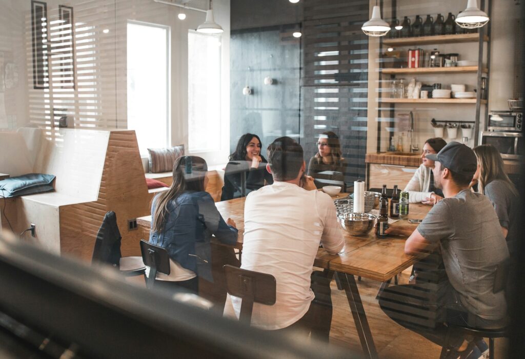 A group of seven people sitting around a wooden table in a modern office kitchen, engaged in conversation.