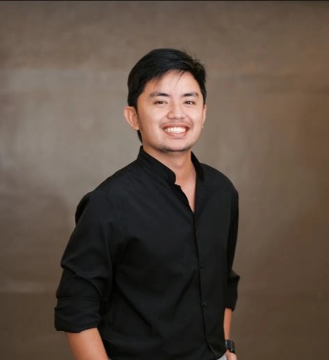 **Alt Text:** A man with dark hair smiling in a black shirt against a neutral brown backdrop.