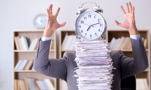 Person in a suit with an alarm clock for a head behind a towering paper stack.