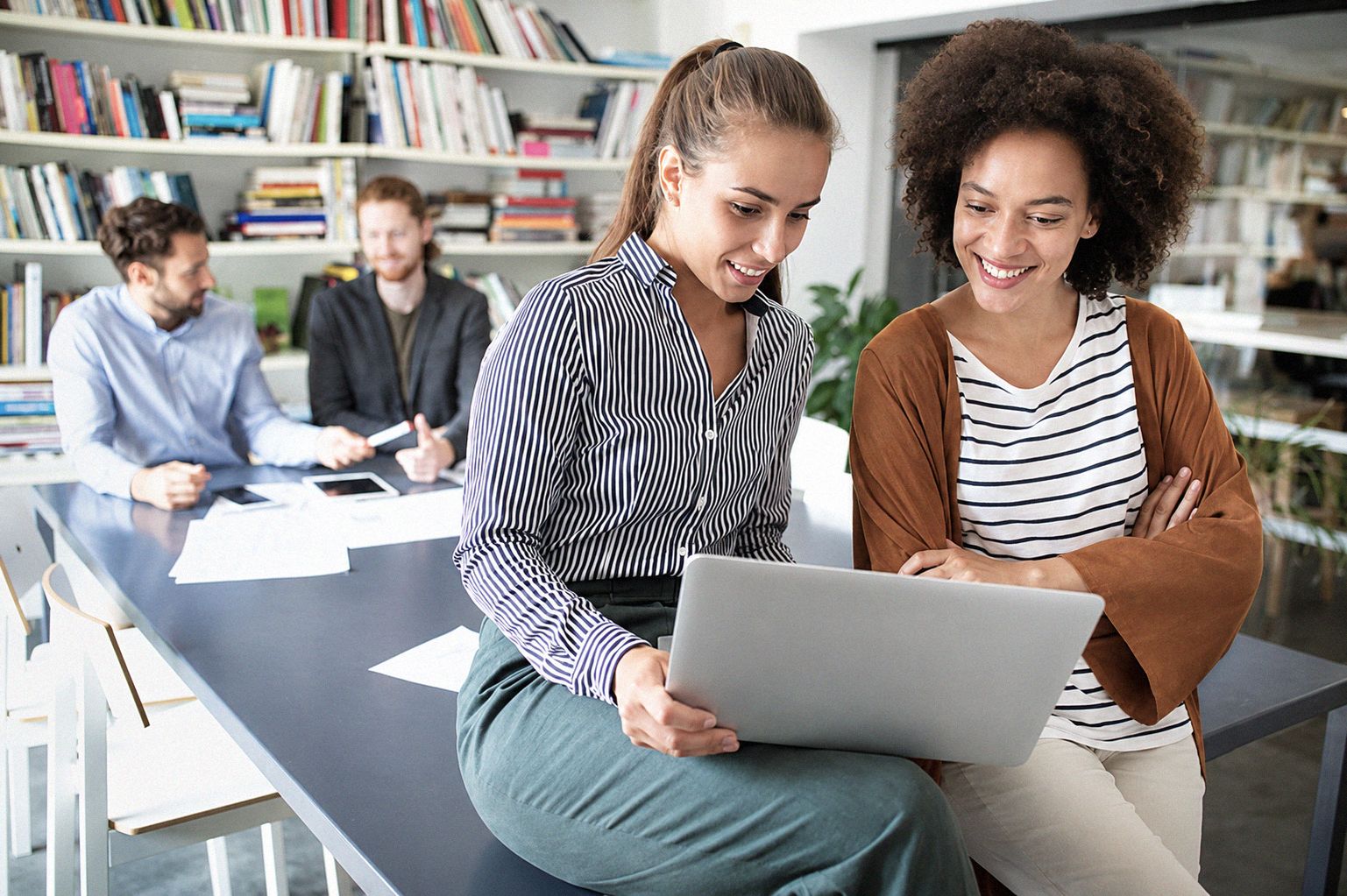 Two smiling women look at a shared laptop while two colleagues talk at a table behind.