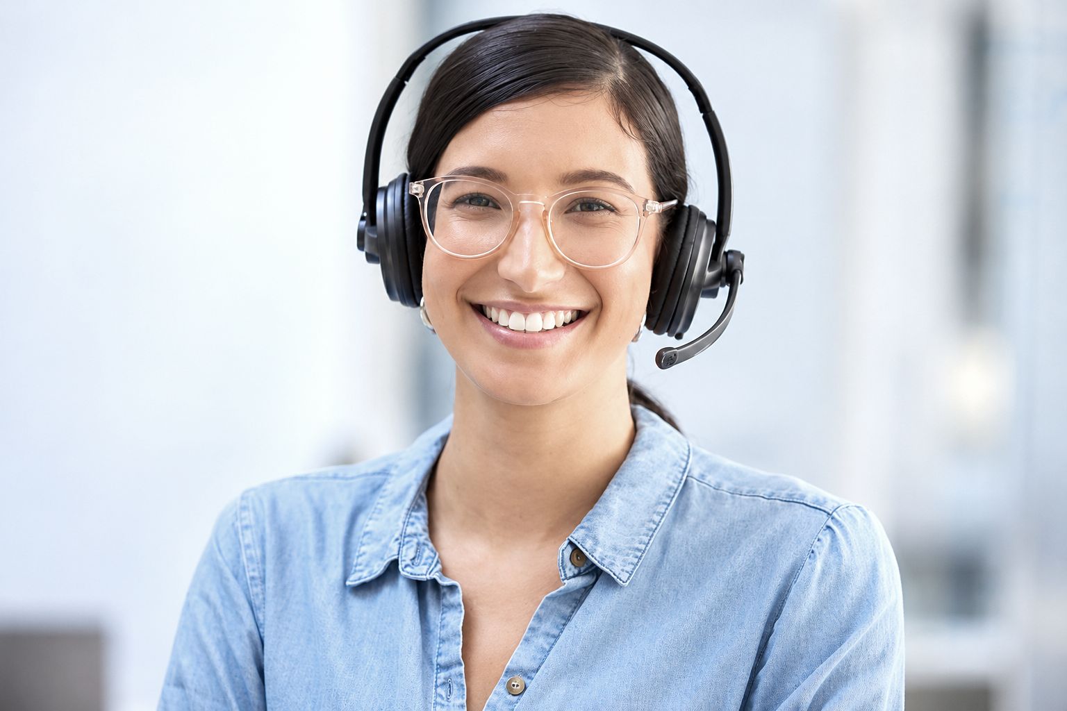 Smiling woman in glasses and a headset wears a light blue denim shirt.