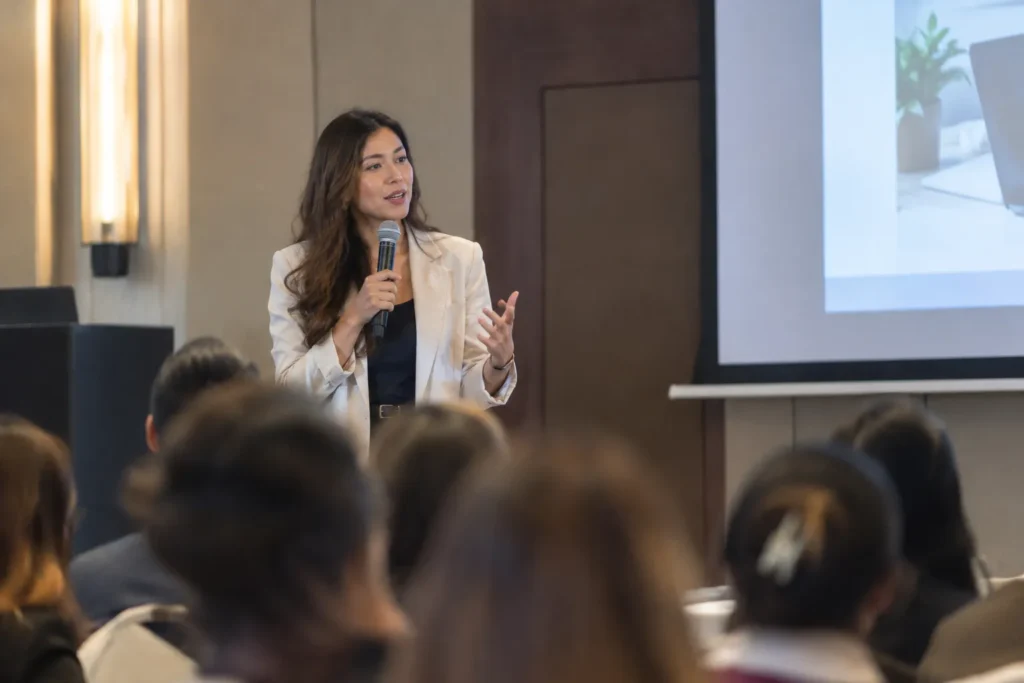 A woman in a beige blazer speaking into a microphone while presenting to an audience at a seminar.
