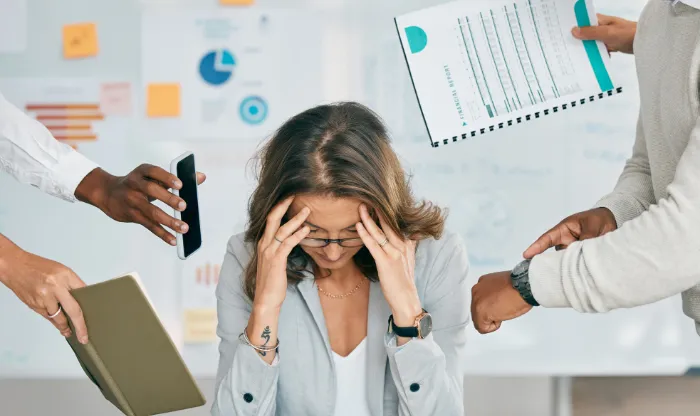An overwhelmed woman holding her head at a desk as colleagues reach toward her with tasks and reports.