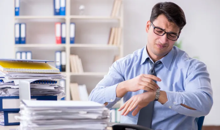 A stressed businessman in a shirt and tie checking his watch beside stacked paperwork on a desk.
