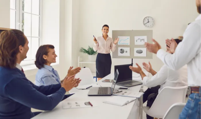 Colleagues clapping at a conference table for a woman presenting chart boards in a bright meeting room.