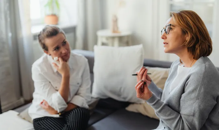 A woman in glasses gesturing while speaking with a colleague sitting on a sofa during a coaching session