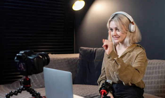 A woman wearing white headphones gesturing toward a laptop and camera setup during a recording session